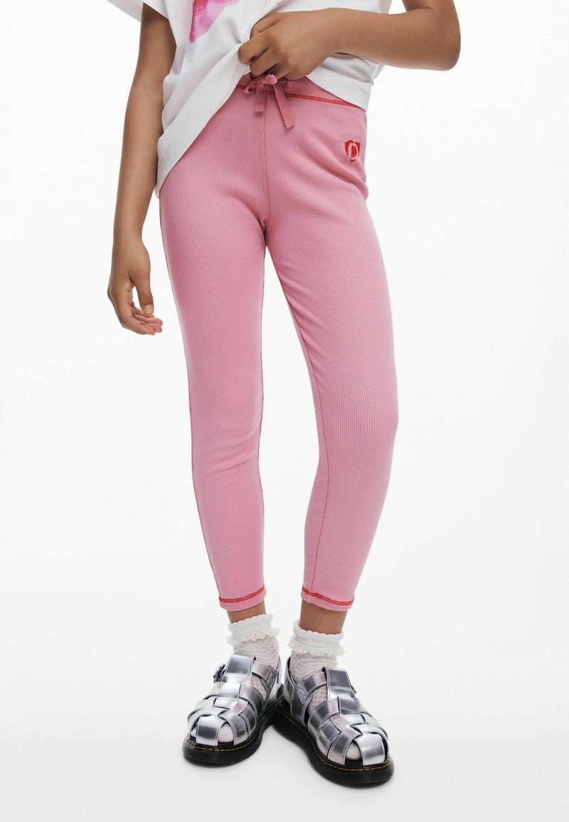 Dijete u ružičastim rebrastim tajicama s crvenim šavovima, bijelim čipkastim čarapama i metalik srebrnim sandalama zatvorenih prstiju, držeći bijelu košulju.