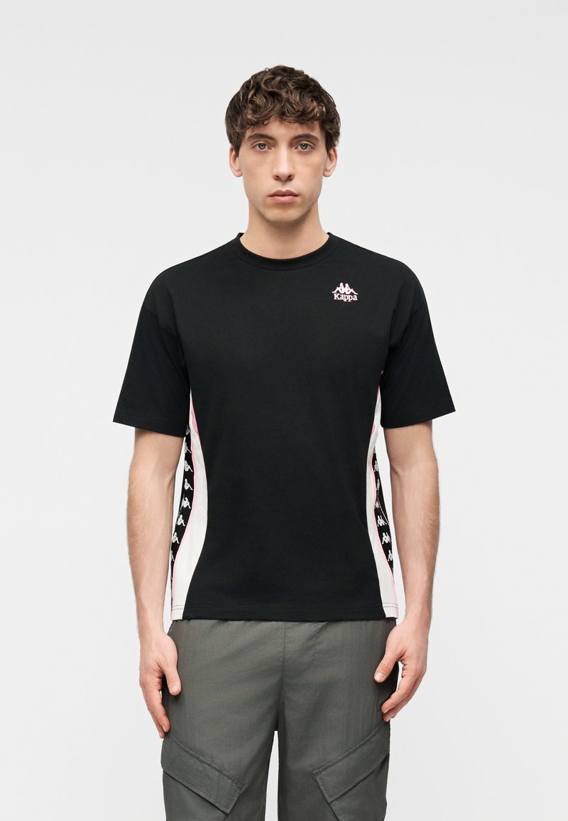 Νεαρός με σγουρά μαλλιά φοράει μαύρο μπλουζάκι Kappa με λευκές και ροζ ρίγες στο πλάι και γκρι cargo παντελόνι, στέκεται μπροστά σε απλό φόντο.