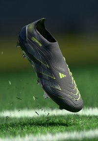 Black and neon yellow Adidas Predator soccer cleat airborne above grass field, with dirt and grass blades flying off the sole studs.