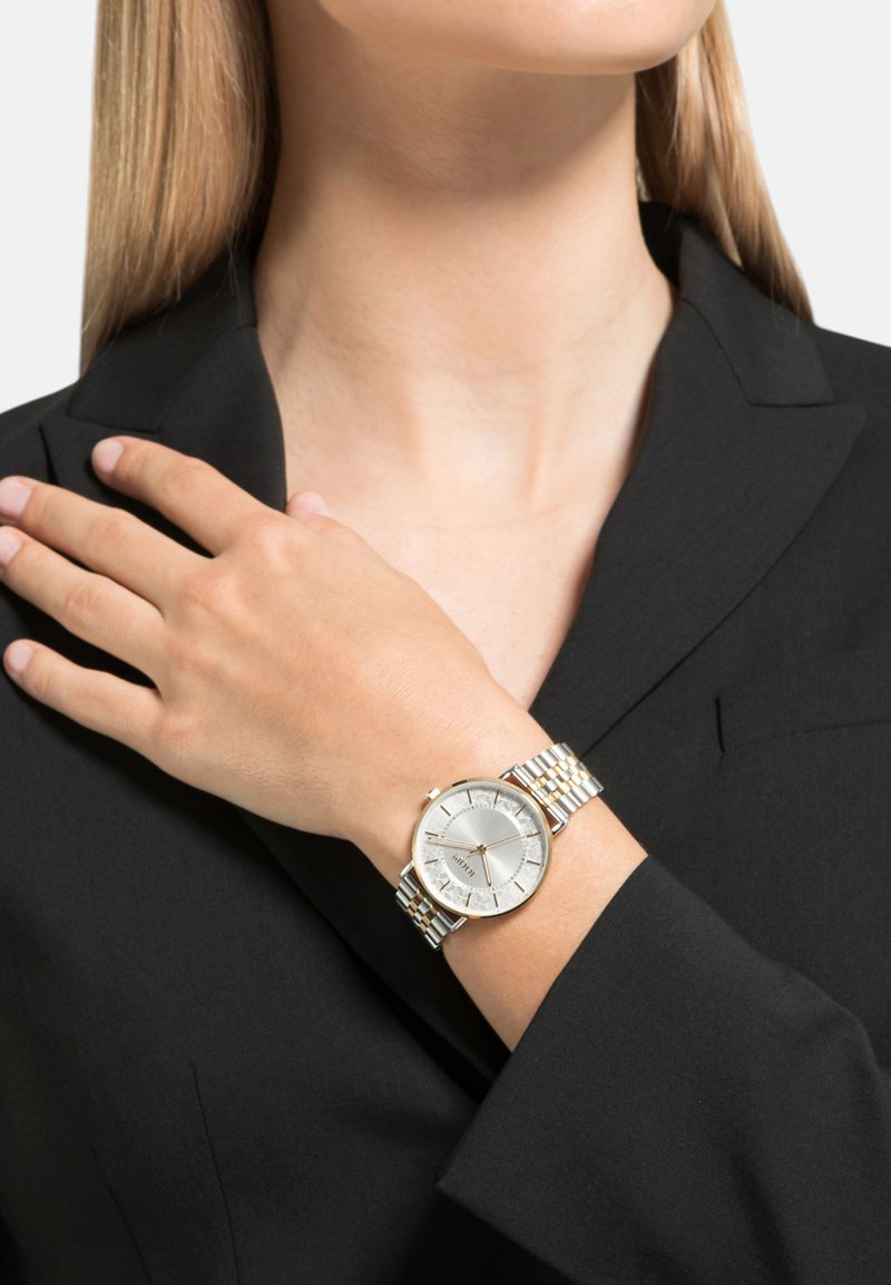 Silberne und goldene Armbanduhr mit rundem Zifferblatt, das ein strukturiertes silbernes Ziffernblatt, ein metallisches Armband und Ziffern aufweist. Am Handgelenk getragen, während es gegen einen schwarzen Blazer angelehnt ist.