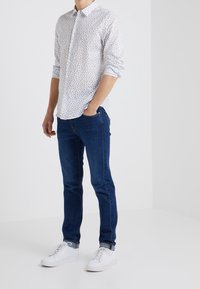 Camicia bianca con piccoli motivi floreali blu, abbinata a jeans blu scuro e sneakers bianche. Design semplice, vestibilità casual.