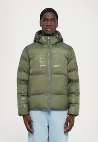 Tânăr îmbrăcat într-o jachetă puffer verde măsliniu cu glugă și modele discrete de logo, purtând o pereche de blugi albastru deschis, stând cu spatele la un fundal simplu.