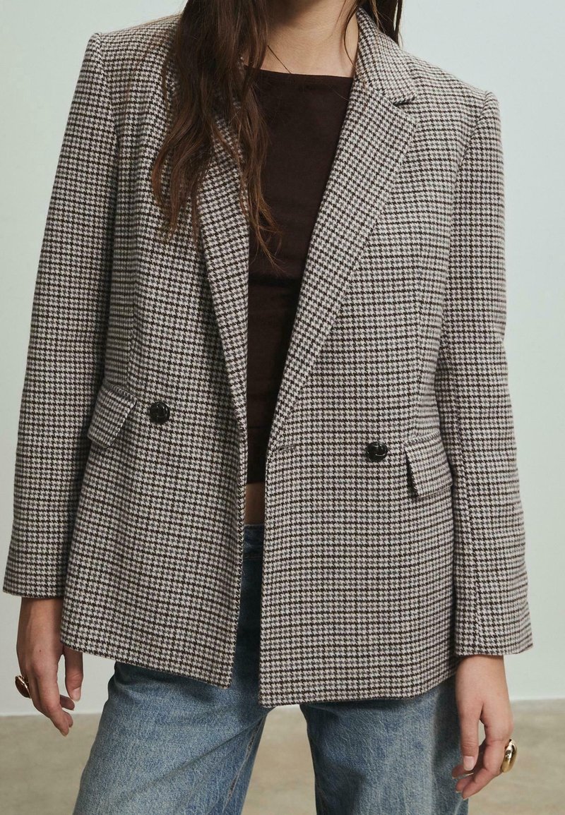 Femme portant un blazer pied-de-poule marron et blanc sur un haut marron foncé et un jean bleu, debout, les bras relâchés le long du corps.