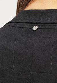 Svart tygskjorta med en strukturerad krage, silverdetaljer på baksidan och en texturerad yta med synliga sömmar.