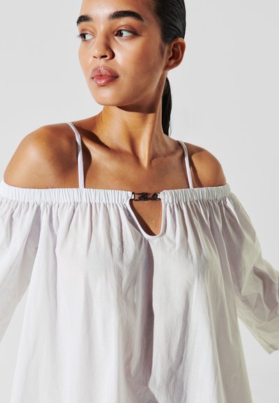 Blouse blanche tombant sur les épaules en tissu léger, avec un col froncé, une découpe en forme de trou de serrure et un accent métallique à l'avant.
