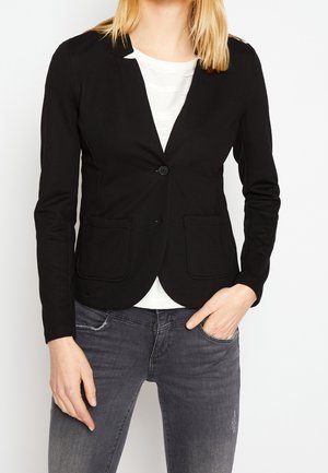 Kobieta ubrana w dopasowaną czarną marynarkę z dwoma przednimi kieszeniami, na białej bluzce i wyblakłych szarych dżinsach, z jedną ręką w kieszeni.