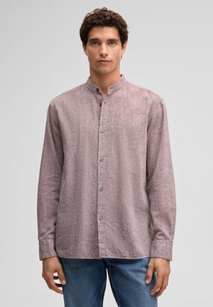 Man met kort krullend haar, die een lichtpaars, geknoopt overhemd met lange mouwen en een blauwe spijkerbroek draagt, staand tegen een effen achtergrond.