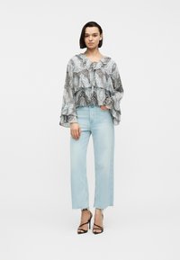 Femme portant un chemisier transparent à motifs avec des volants, un jean bleu clair court aux ourlets effilochés, et des talons hauts noirs à brides, debout.