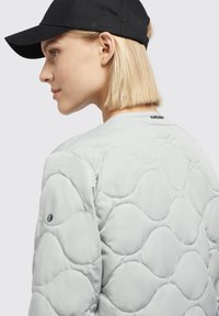 Lichtgrijze gewatteerde jas met een gestructureerd golfpatroon, voorzien van een zwart logo op de mouw en een eenvoudige zwarte pet die achterstevoren wordt gedragen.