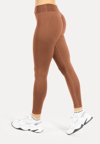 Braune Leggings mit hohem Bund, figurbetontem Design, glatter Textur und Nahtdetails, kombiniert mit weißen Sportschuhen.