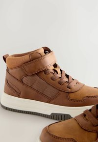 Brązowe skórzane sneakersy za kostkę z teksturowanym bocznym panelem, wyściełanym kołnierzem, regulowanym paskiem i białą gumową podeszwą.