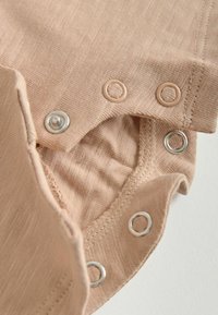 Tissu beige avec boutons-pression en métal et plastique partiellement défaits, montrant les coutures et les détails du matériau texturé.