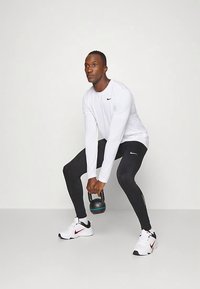 Mies, joka pukeutunut valkoiseen pitkähihaiseen paitaan ja mustiin leggingsseihin, nostaa kahvakuulaa kyykkyasennossa yksinkertaista taustaa vasten.