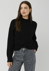 Pull en maille noir surdimensionné avec un col haut et des poignets côtelés, associé à un jean gris orné d'un motif léopard noir.