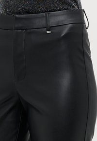 Schwarze Kunstlederhose mit figurbetontem Design, glatter Textur, einer Fronttasche und einem kleinen metallischen Akzent am Bund.