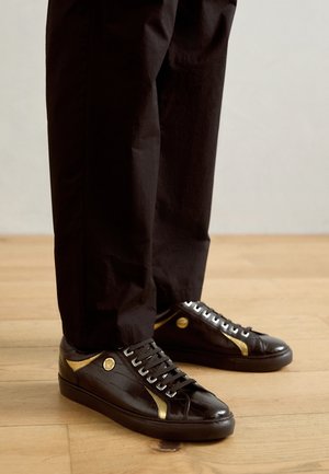 Piernas con pantalones negros y zapatillas de cuero negro con acentos dorados, de pie sobre un suelo de madera clara.