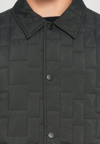 Veste matelassée noire avec motif de points carrés, col pointu et boutons-pression noirs sur la fermeture avant.