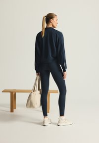 Marineblå atletisk sweatshirt og leggings med en glat tekstur, parret med en crèmefarvet taske. Modellen står på en træbænk mod en lys baggrund.