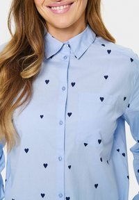 Camicia azzurro chiaro con colletto, decorata con motivi a cuore navy, taschino sul petto e una texture di cotone liscia.