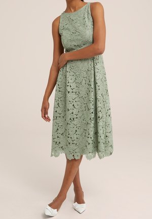 Sukienka midi bez rękawów z kwiatowym wzorem z koronki w jasnym zielonym kolorze, noszona z białymi szpilkami o spiczastym nosku, modelka stoi z założonymi skrzyżowanymi ramionami.