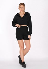 Felpa nera corta con zip frontale, maniche lunghe, abbinata a pantaloni shorts neri a vita alta. Il modello indossa scarpe da ginnastica nere. Tessuto dalla superficie liscia.