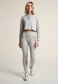 Lichtgrijze cropped hoodie met een trekkoord en logo, gecombineerd met bijpassende high-waist leggings. Witte branding op de leggings, casual sneakers.