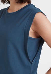 Woman wearing a sleeveless blue top with wide armholes, showing a fitted inner layer underneath, against a white background.