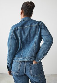 Keskmise pesu denimjakki struktureeritud lõikega, ees yoke õmblustega ja nööbiga kinnitusega, koos sobivates denimpükste komplektiga.