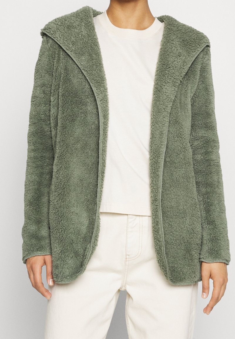 Green fleece jacket with a hood, featuring a soft texture and open front design, worn over a light t-shirt and light-colored pants.
