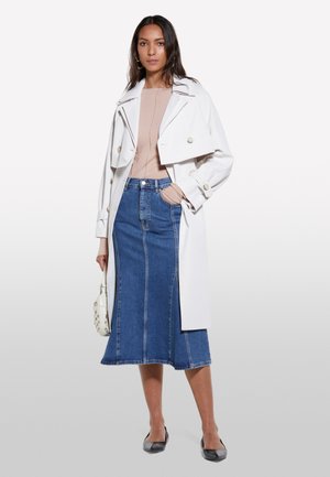 White trench coat with double-breasted buttons, paired with a light pink top and blue denim midi skirt. Black flats and small white handbag complete the look.