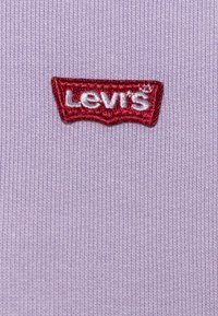 Un tessuto lavanda con una texture a coste presenta un logo Levi's ricamato in rosso su un badge curvato. Il design enfatizza l'identità del marchio.