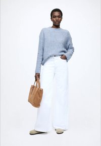 Šviesiai mėlynas megztinis, balti plataus kirpimo džinsai, ruda zomšinė rankinė ir smėlio spalvos batai. Modelis pozuoja su viena ranka kišenėje, atsipalaidavusioje pozoje.