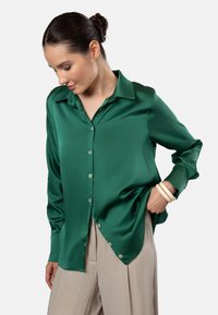 Green satin button-up shirt with a pointed collar, long sleeves, and a curved hem. Features light button accents and smooth texture.