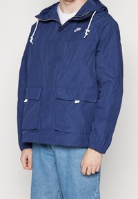 Giacca a vento blu navy con cappuccio, caratterizzata da una finitura testurizzata, due tasche frontali e dettagli bianchi sui lacci. Logo sul petto.