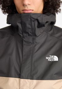 Juodas ir smėlio spalvos švarkas su aukštu apykakle, pagamintas iš tekstūruotos medžiagos; kairėje krūtinės pusėje yra balta "The North Face" logo.