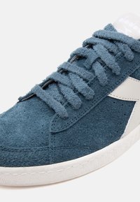 Blue suede sneaker featuring a textured upper, flat laces, and white rubber sole with a smooth finish and side logo detail.