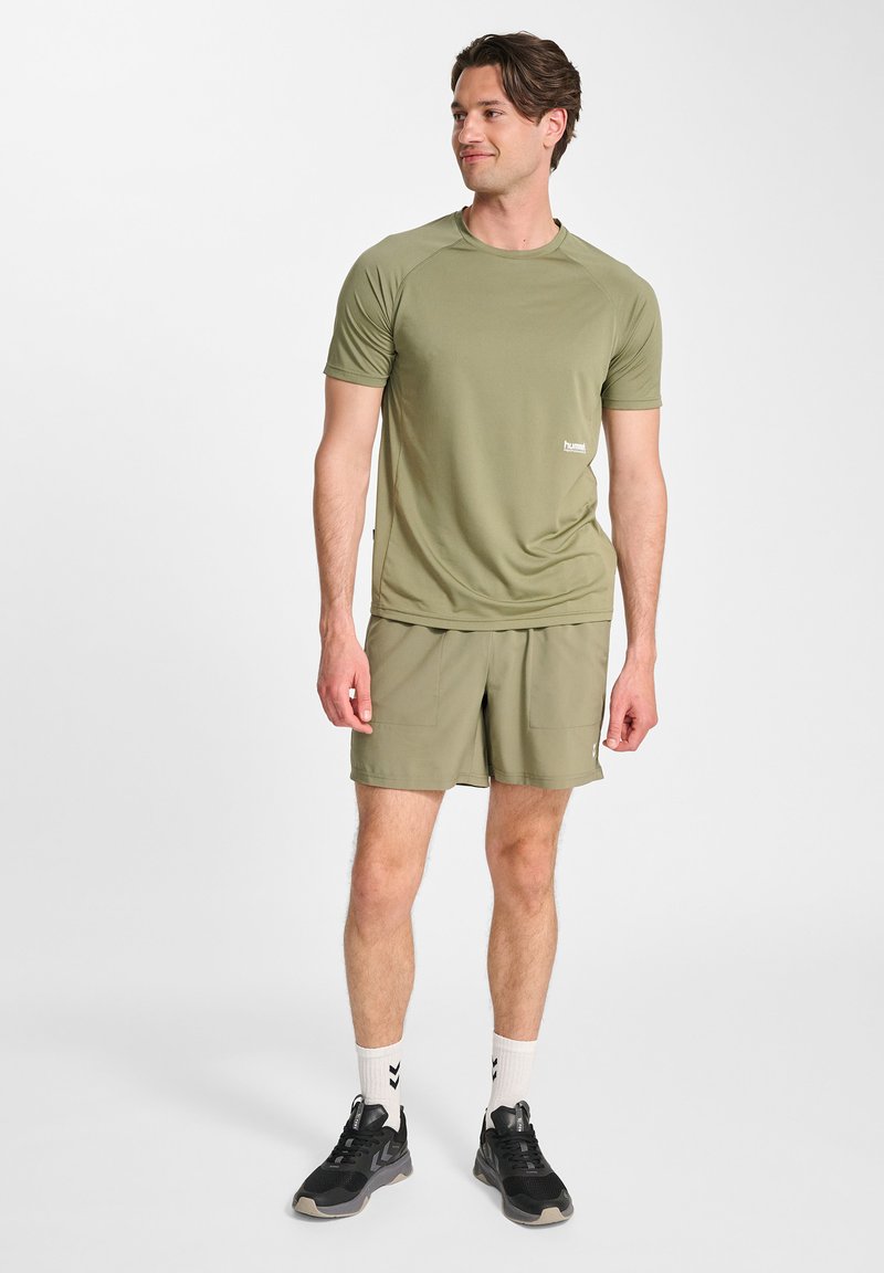 Olivgrön idrottströja med korta ärmar, matchad med kortbyxor. Svarta sneakers med vita strumpor som har ett chevronmönster.