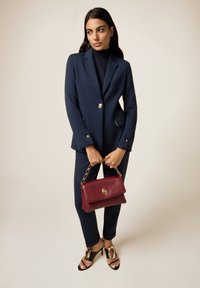 Costume bleu marine avec une veste sur mesure et un pantalon slim, associé à un sac à main bordeaux et des sandales noires avec des accents dorés. Texture lisse.