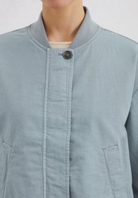 Light blue jacket made of soft fabric, featuring a standing collar, single button closure, and two front pockets. Smooth texture with subtle stitching.
