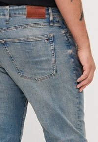 Világoskék farmernadrág, kopott hatással, amelyen a derékrészen a "TOMMY JEANS" felirattal ellátott barna bőrpárna és diszkrét márkaelemek találhatók.