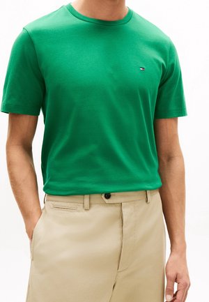 Man wearing a plain green short-sleeve t-shirt and beige pants with one hand in pocket against a white background.