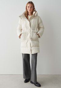 Manteau long et bouffant de couleur crème avec une capuche, des boutons-pression et deux poches avant. Tissu texturé avec un design chaud et matelassé.