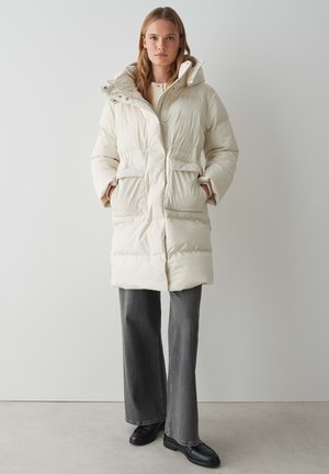 Long, puffy cream-colored coat with a hood, snap buttons, and two front pockets. Textured fabric with a warm, quilted design.