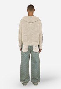 Υπερμεγέθη μπεζ πλεκτή κουκούλα με ριγωτή υφή, σε συνδυασμό με φαρδιά ανοιχτό πράσινα παντελόνια που διαθέτουν επαναλαμβανόμενο σχέδιο και αντίθετες τσέπες.