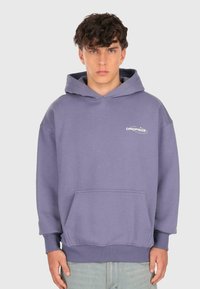 Junger Mann mit lockigem Haar, der einen lavendelfarbenen Hoodie mit Fronttasche und dem Logo "DROPSIZE" trägt, steht vor einem schlichten hellen Hintergrund.