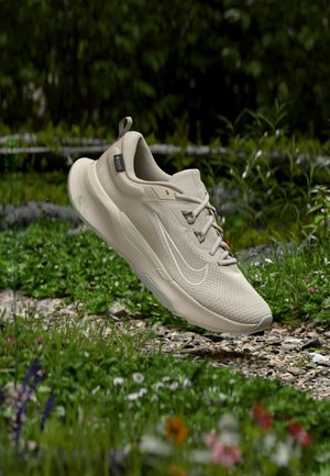 Scarpa da ginnastica atletica beige con tomaia testurizzata e logo, sospesa sopra un'area erbosa con piccoli fiori e un sentiero di ghiaia, ambiente esterno.