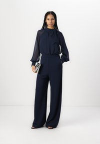 Mørkeblå jumpsuit med høy hals og gjennomsiktige langermede ermer; har en vide ben-design og glatt stoff. Kompletteres med en metallisk clutch.
