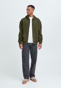 Jonge man staat casual gekleed in een olijfgroen hoodie, wit T-shirt en donkere spijkerbroek, met een neutrale achtergrond.