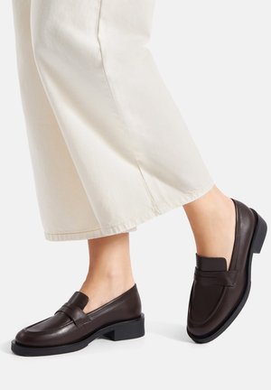 Braune Ledermokassins mit niedrigem Blockabsatz, verziert mit einer gesteppte Details und einem dekorativen Riemen. Kombiniert mit cremefarbenen Hosen mit weitem Bein.