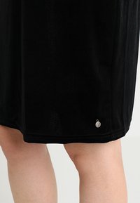 Ourlet de jupe en velours noir avec petit pendentif rond argenté sur les jambes d'une personne, sur fond uni clair.